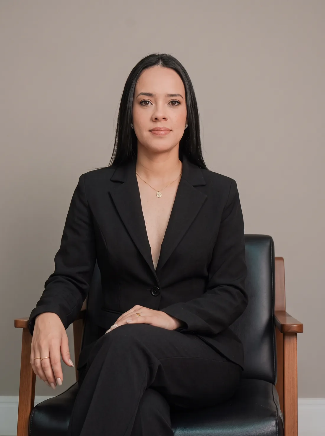 Dra. Jordana Sanseverino, advogada, de blazer preto e cabelo longo, sentada em uma poltrona de couro preto.